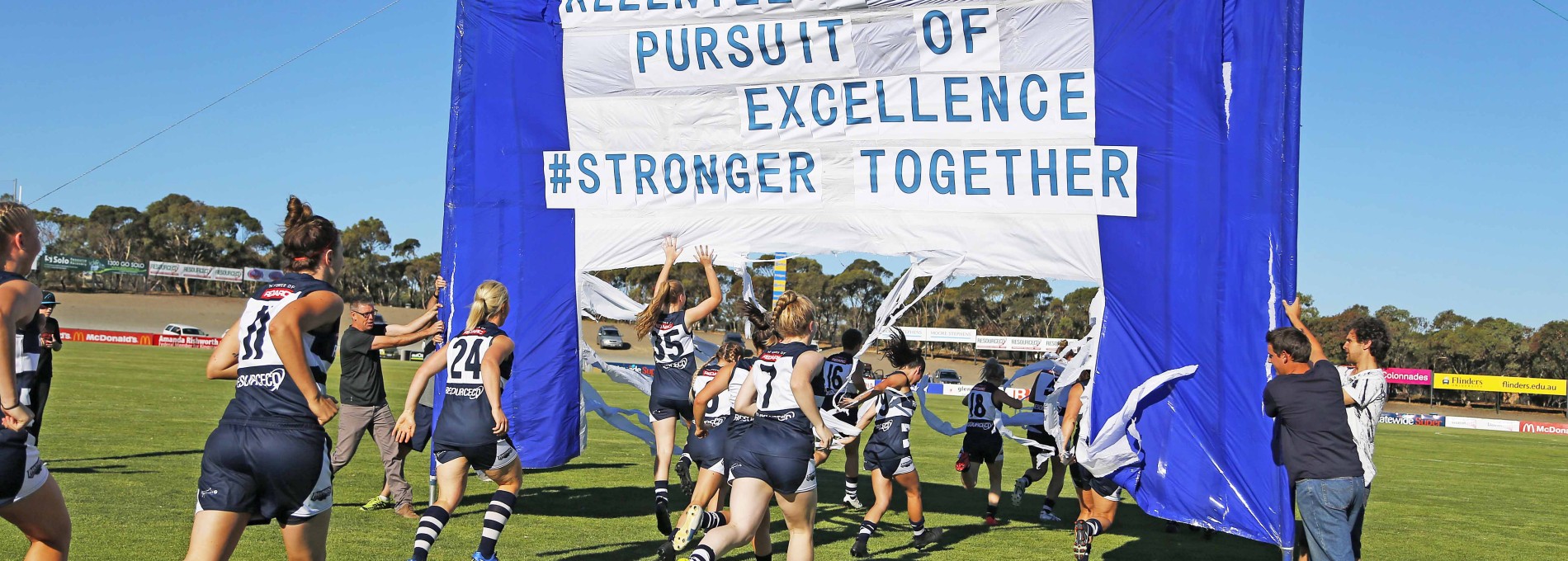 A Young Female AFL Team running on field through Team Flag